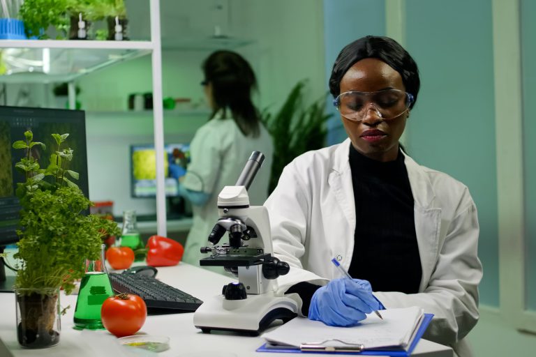 Biologist researcher scientist looking at leaf sample under medical microscope while writing biological expertise on notepad. Chemist examining biological discovery working in microbiology lab